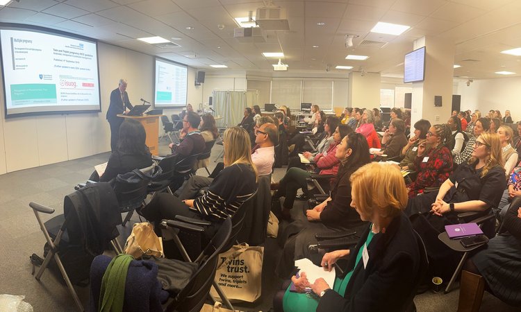 A wide image of a room of people seated lecture style looking at a presentation on the left of image