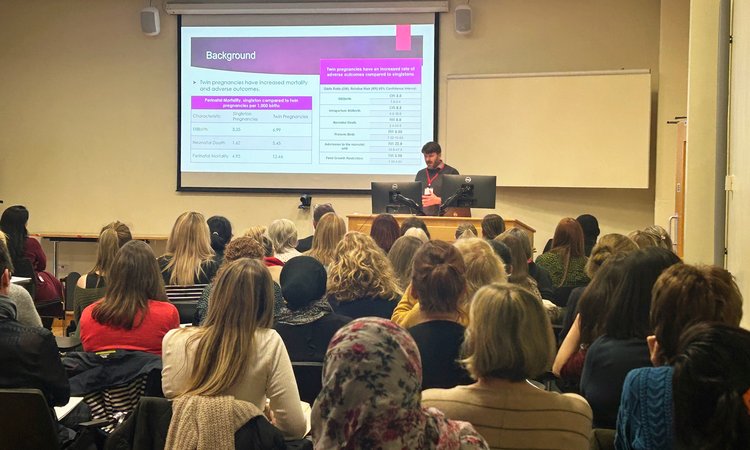 A room of people seated lecture style watching a presentation at the front, taken from behind