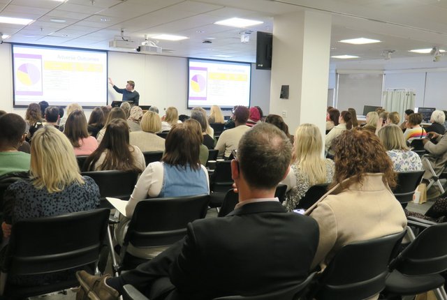 A room of people sat lecture style watching a presentation at the front, taken from the back