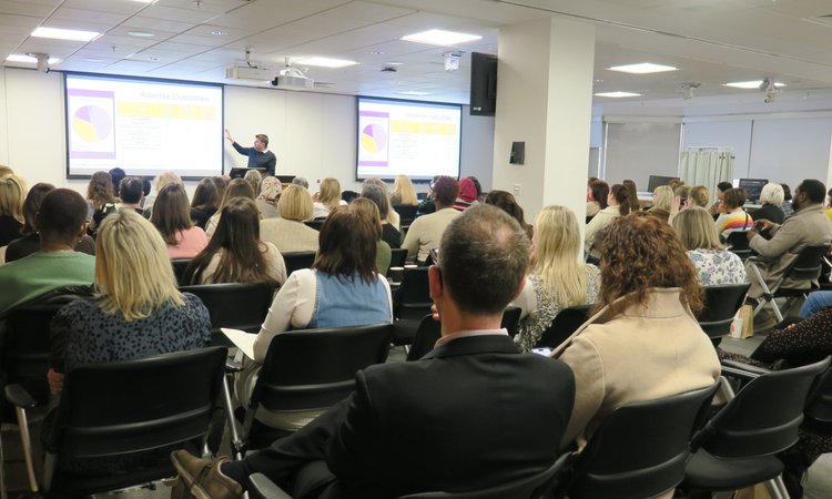 A room of people sat lecture style watching a presentation at the front, taken from the back