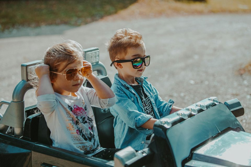 A young boy and girl wearing sunglasses drive a toy car