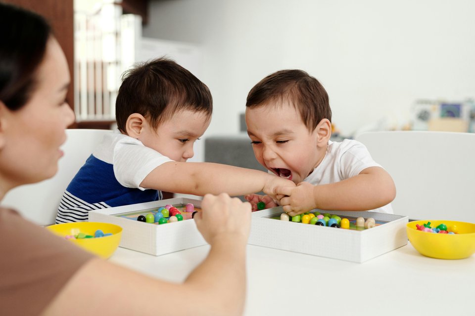 Two young boys sat at a table playing as one tries to bite the hand of the other