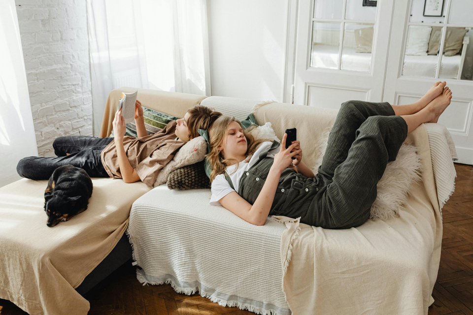 Two teenagers lie on a sofa, one on their phone and one reading a book