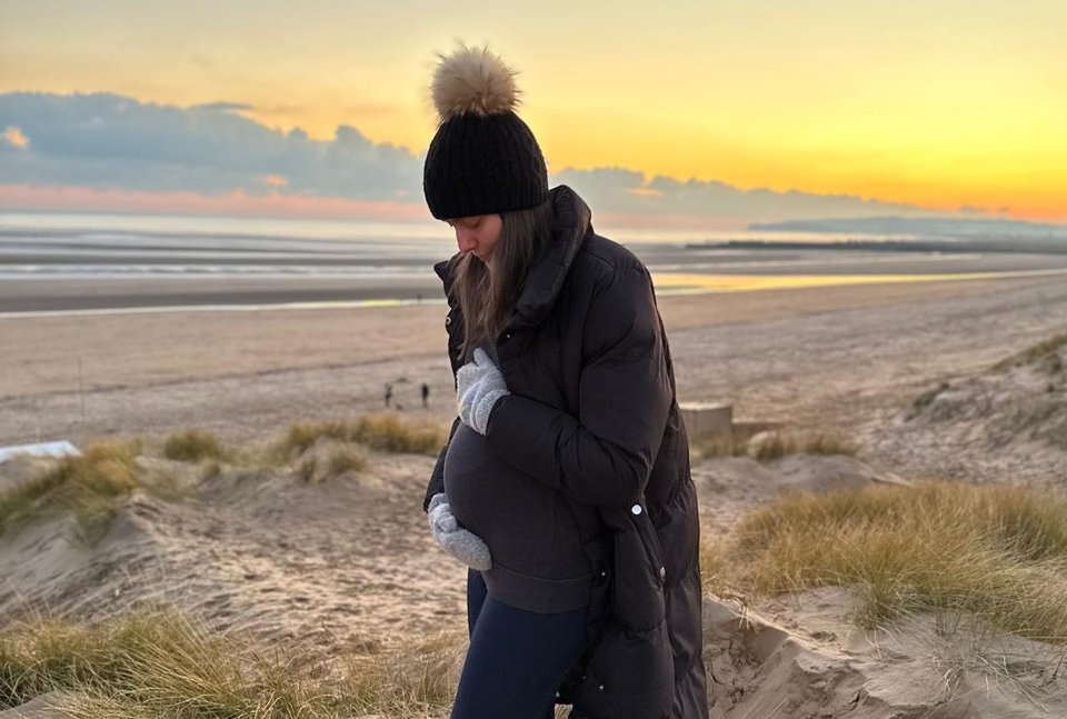 A pregnant woman stands in front of a sunset cradling her baby bump