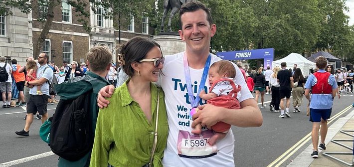 A man and a woman pose together holding a baby. The man has a running finishers medal and number on