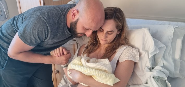 A woman sits in a hospital bed cradling a baby as a man leans over and holds her hand