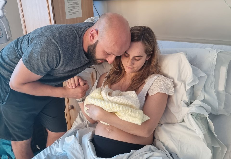 A woman sits in a hospital bed cradling a baby as a man leans over and holds her hand