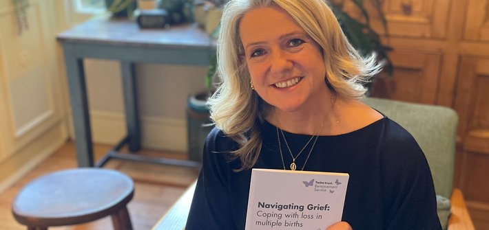 A woman sits smiling at the camera holding a Navigating Grief booklet