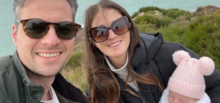A man and woman pose holding a baby with a cliff head and sea in the background