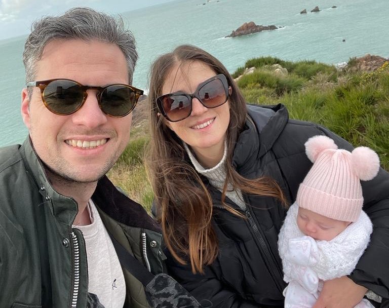 A man and woman pose holding a baby with a cliff head and sea in the background