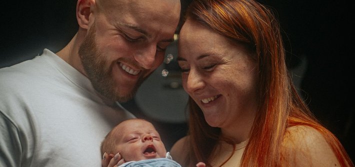 A man and woman stand together holding a baby