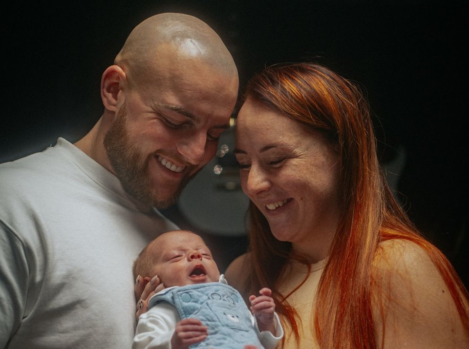 A man and woman stand together holding a baby