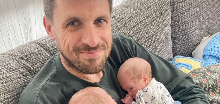 A man sits on a sofa holding two babies against his chest