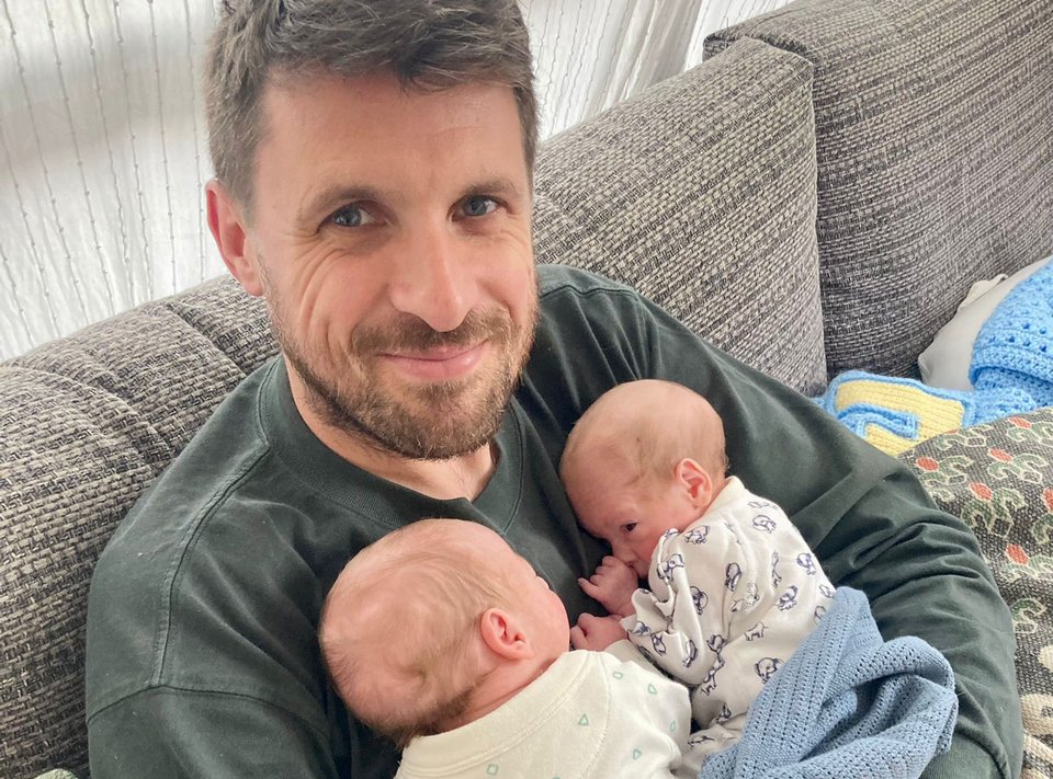A man sits on a sofa holding two babies against his chest