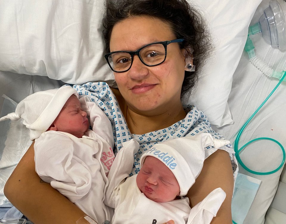A woman in a hospital gown lies on a bed holding two babies