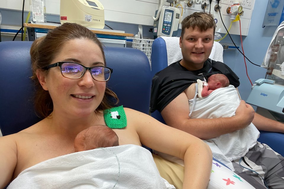 A man and woman sit on hospital chairs each with a baby on their chest