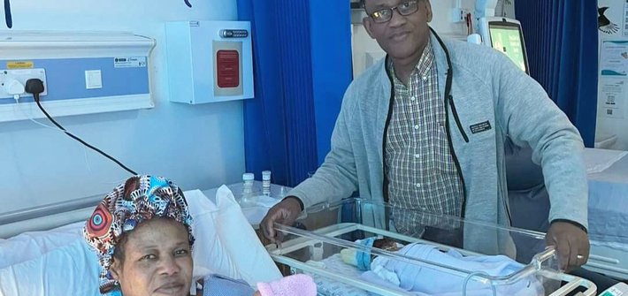 A woman lies in a hospital bed holding a baby as a man stands next to a cot