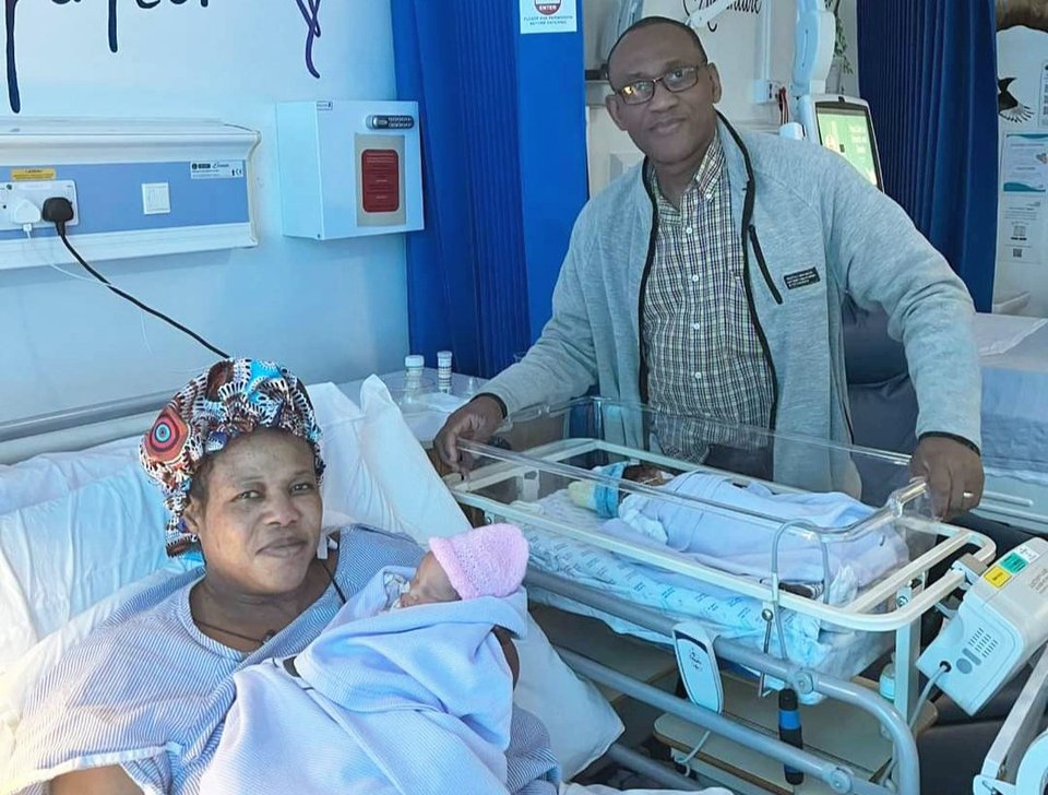 A woman lies in a hospital bed holding a baby as a man stands next to a cot