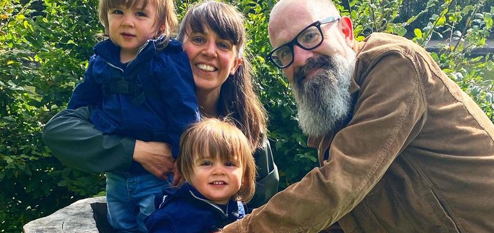 A man and woman stand next to twin children sitting on a tree stump