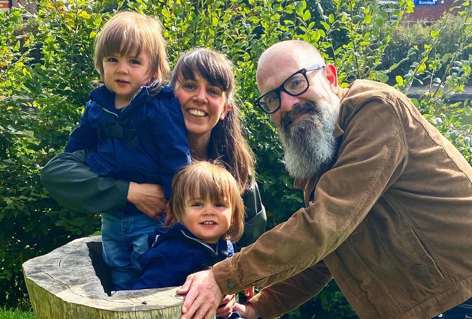 A man and woman stand next to twin children sitting on a tree stump