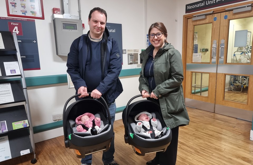 A man and a woman each hold a baby carrier as they prepare to leave hospital