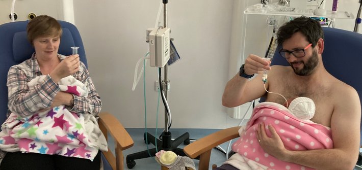 A man and a woman sit on hospital chairs each with a baby in their arms