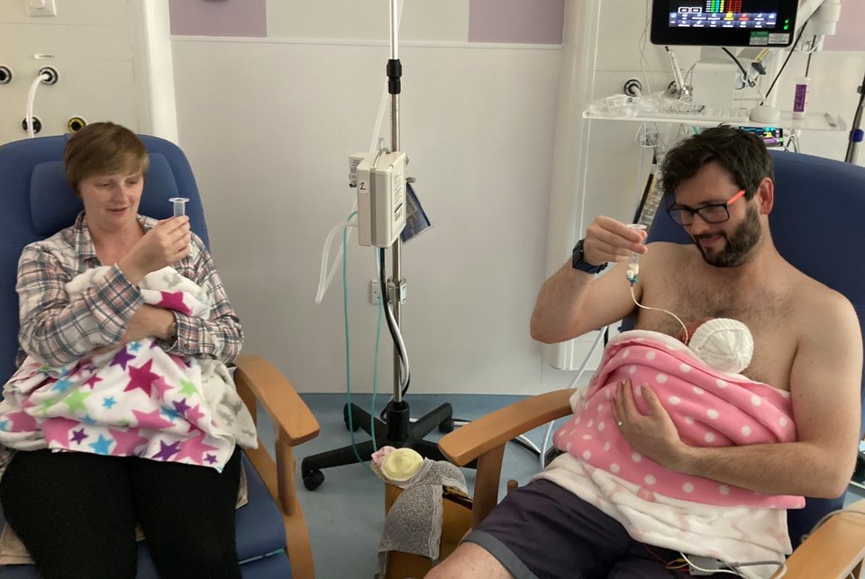 A man and a woman sit on hospital chairs each with a baby in their arms