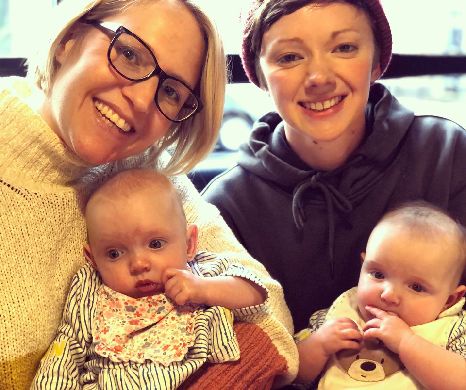 Two women sit smiling holding twin babies
