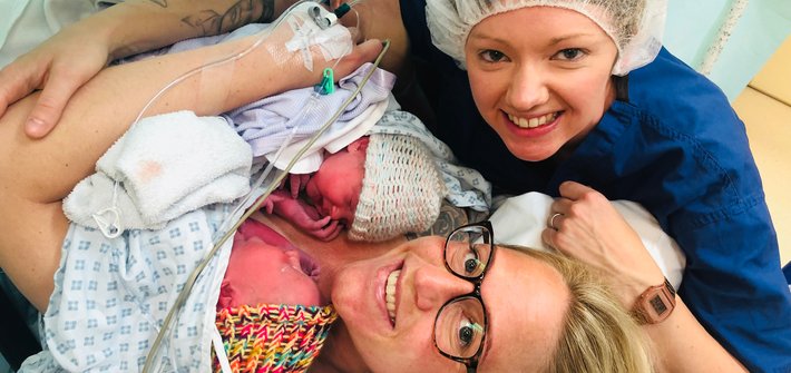 A woman in a hospital gown lies holding two babies as another woman sits with her arm around them