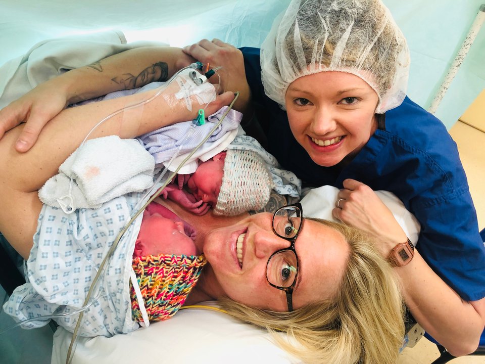 A woman in a hospital gown lies holding two babies as another woman sits with her arm around them