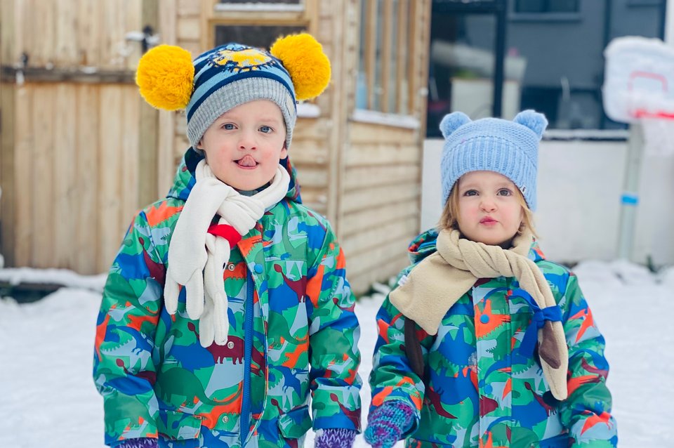 Twins dressed in matching snow suits stand in a snow covered garden