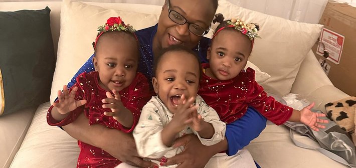 A woman sits on a sofa with triplets on her lap