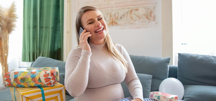 A pregnant woman sits on a sofa surrounded by presents talking on the phone