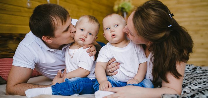 Twin babies sit on a bed with a mother and father either side kissing their cheeks