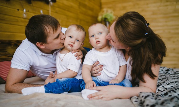 Twin babies sit on a bed with a mother and father either side kissing their cheeks