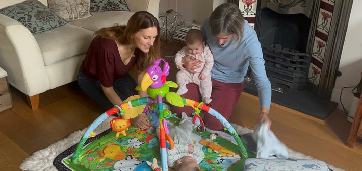 Two women kneel on the floor, one holding a baby, as they tend to a baby lying on a mat