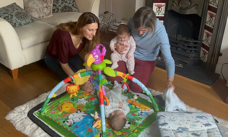 Two women kneel on the floor, one holding a baby, as they tend to a baby lying on a mat