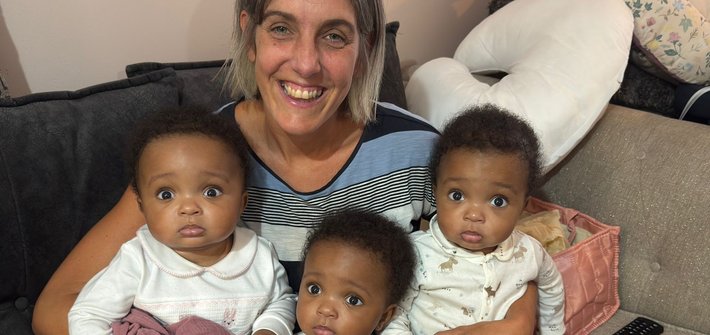 A woman sits on a sofa with young triplets on her lap