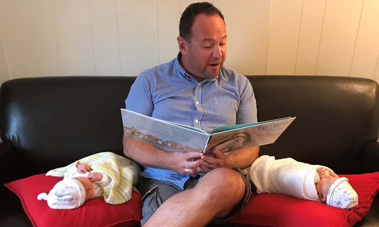A man sits on a sofa reading a book to twin babies lying on pillows either side of him