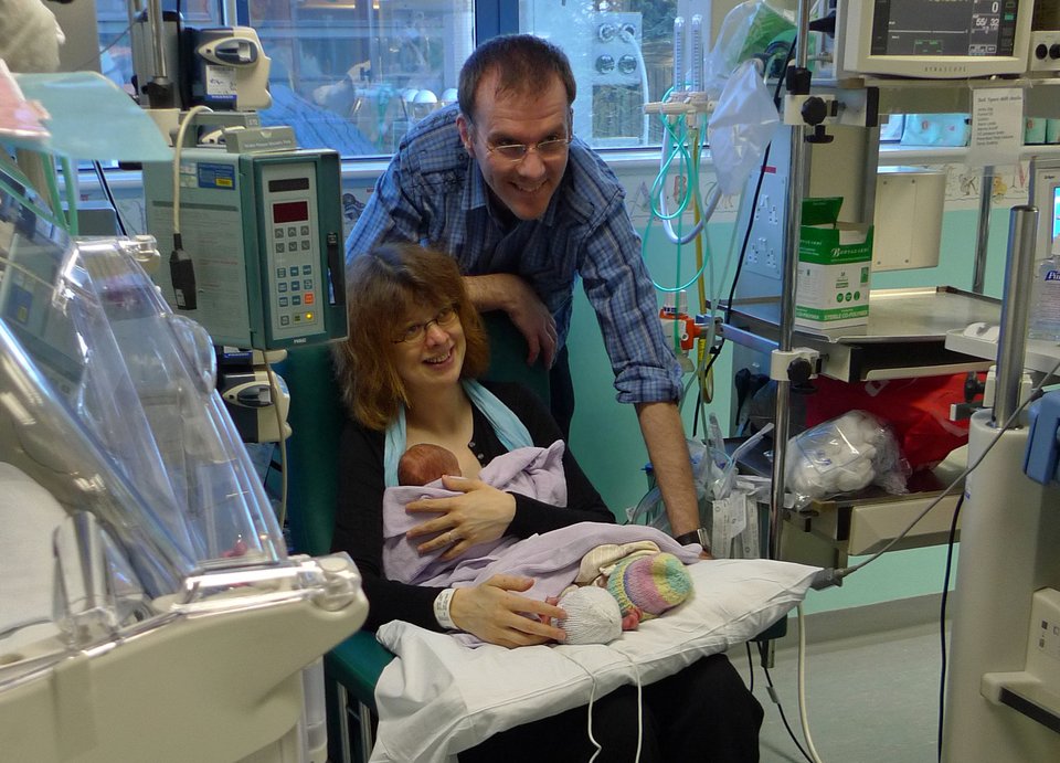 A woman in a hospital chair holding three babies with a man leaning over her shoulder