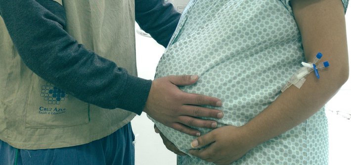 A close-up of a pregnant woman in hospital gown and another person next to her, both cradling the baby bump