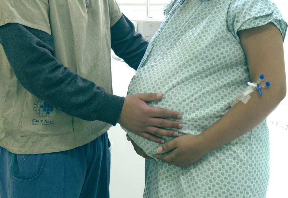 A close-up of a pregnant woman in hospital gown and another person next to her, both cradling the baby bump