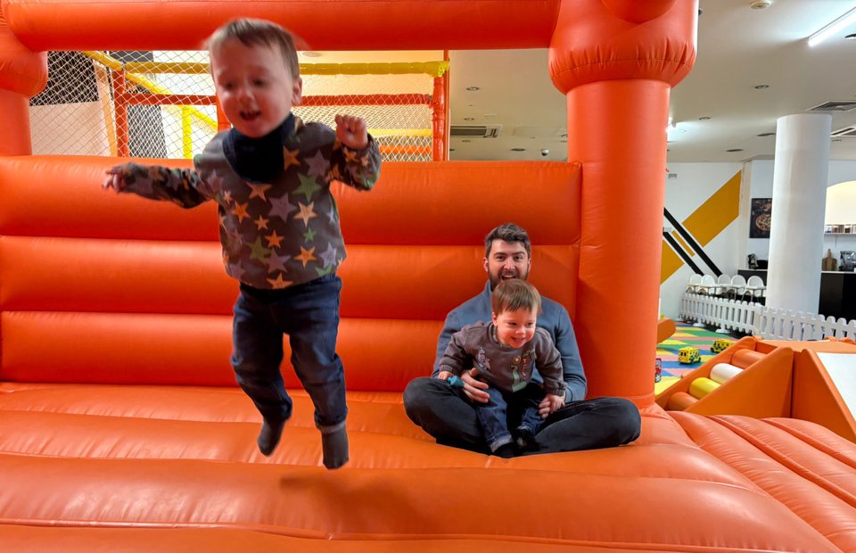 A boy bounces on a trampoline as a man sits with another boy in the background
