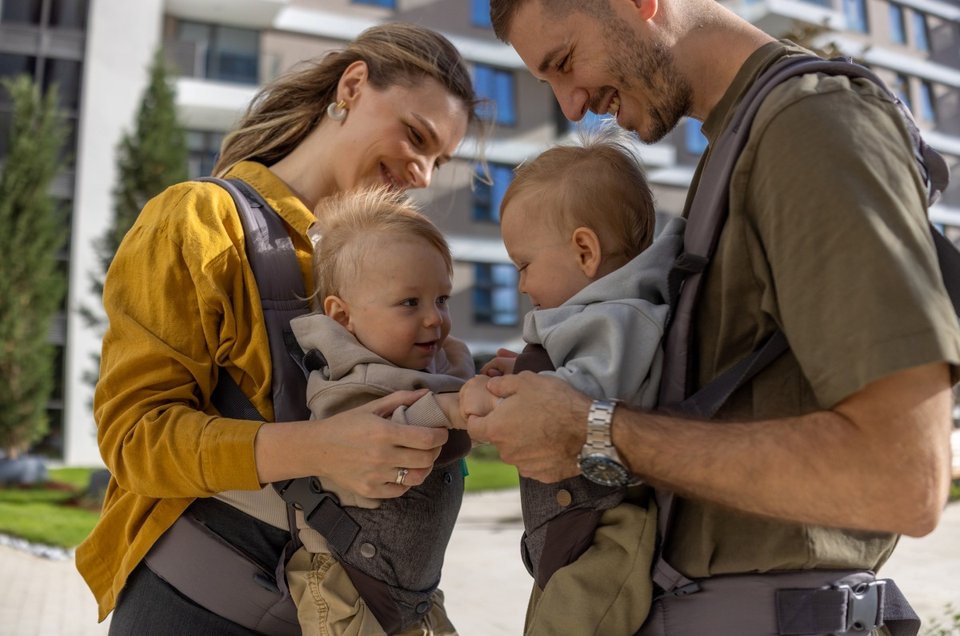 a man and a woman each carrying a twin child in a baby carrier