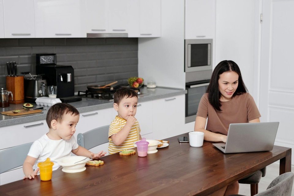 a mother using a laptop in the kitchen with her twin boys eating at the table