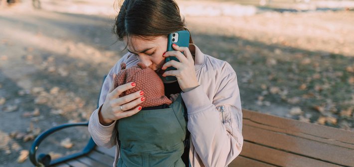 A woman sitting on a bench talking on the phone as she kisses the head of a baby in a carrier