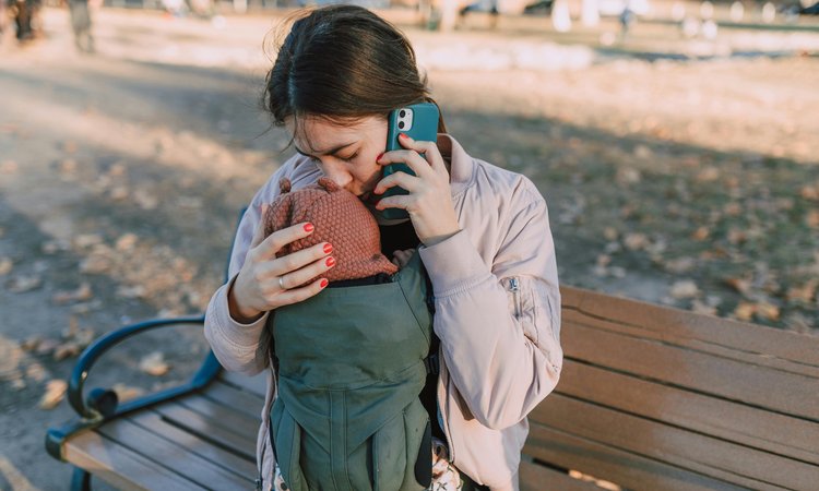 A woman sitting on a bench talking on the phone as she kisses the head of a baby in a carrier
