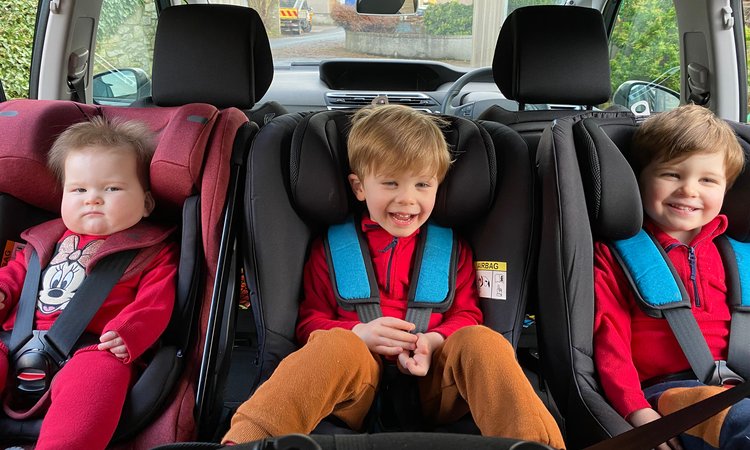 Three children sit in car seats in the back of a car