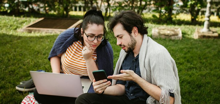 A man and woman sit next to each other on grass as the man shows the woman something on a smartphone