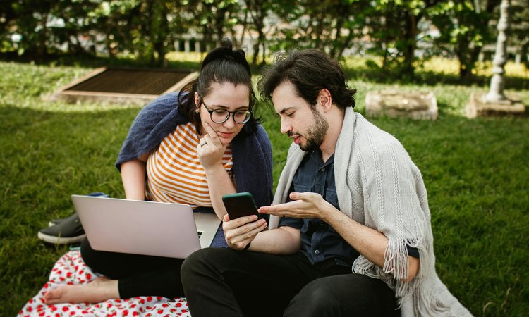 A man and woman sit next to each other on grass as the man shows the woman something on a smartphone